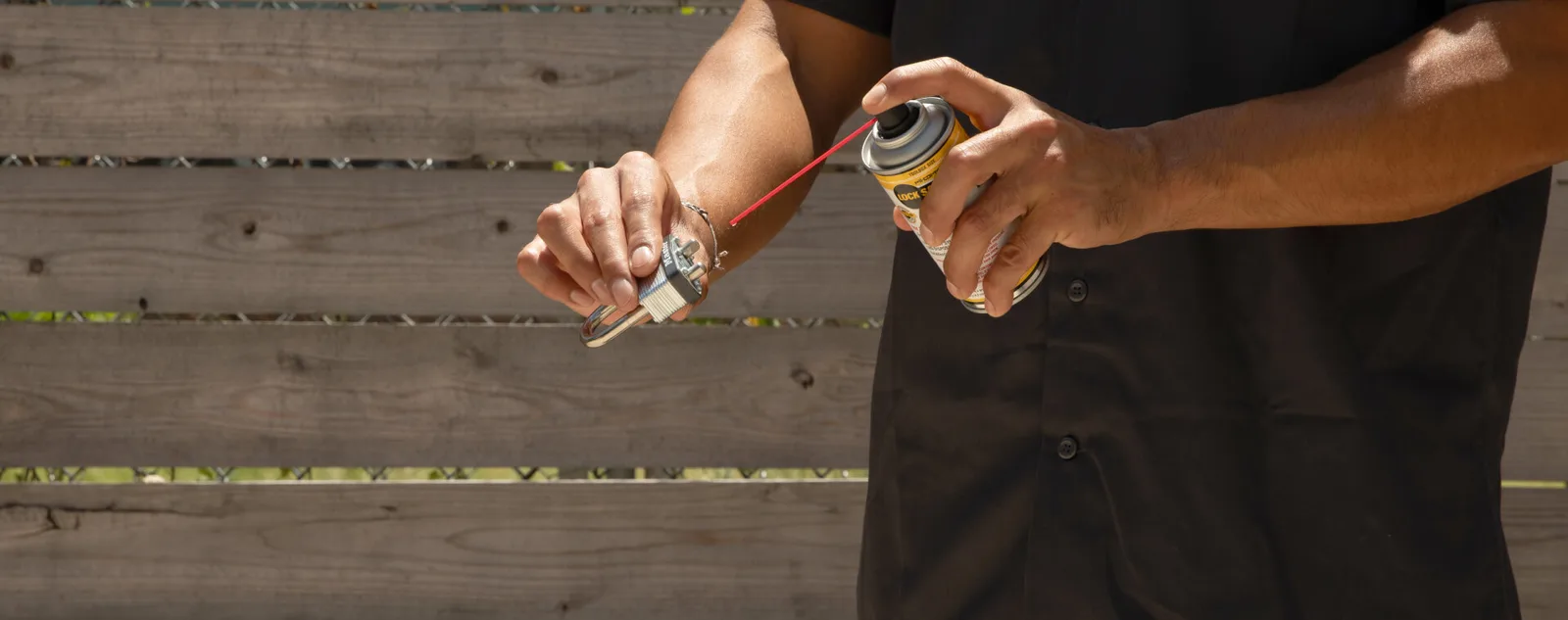 A man in a black work shirt spraying Lock Saver through the red straw into a padlock, in front of a wooden fence