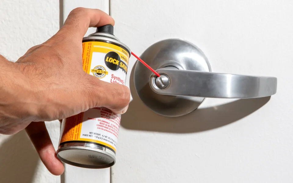 A hand sprays Lock Saver through its red extension straw into the keyway of a brushed steel lever door handle